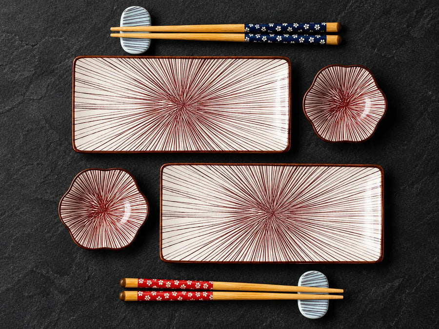 A flat-lay view of a Japanese-style dinnerware set on a dark textured slate background. The set includes two rectangular ceramic plates with a red radial starburst pattern on a cream background, two matching small flower-shaped bowls, and a pair of wooden chopsticks with blue and red floral-patterned handles resting on small white ceramic coasters.