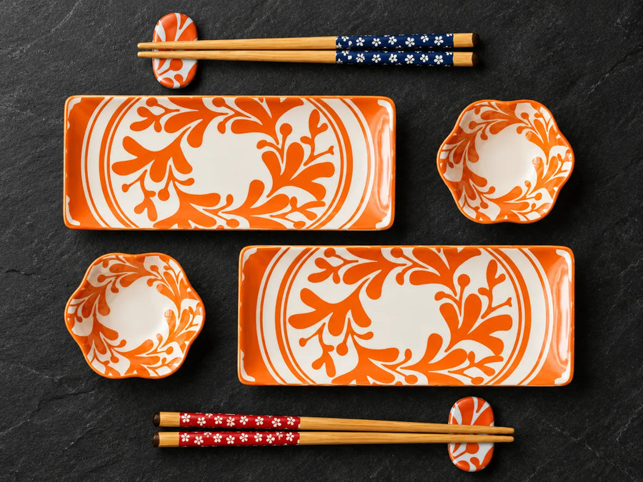 A coordinated dinnerware set arranged on a dark textured surface, including two rectangular plates, two scalloped bowls, and four pairs of chopsticks with decorative handles and matching chopstick rests. The dishes have a white base with bold orange floral scrollwork, and some chopsticks have blue and red patterned handles.