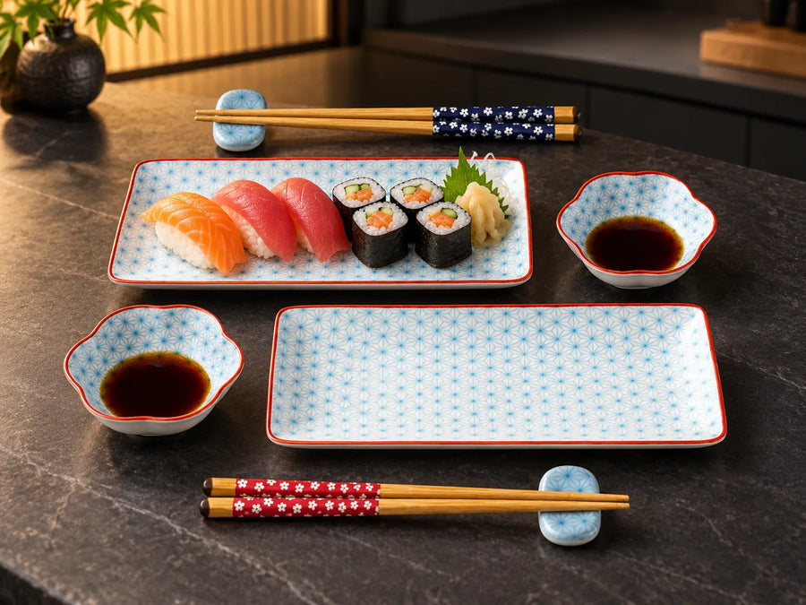 A Japanese-style dinnerware set on a dark stone table, featuring rectangular and scalloped plates with a light blue and white star pattern and red borders, accompanied by matching soy sauce bowls, chopsticks, and small blue coasters. A plate of sushi is arranged on the set.