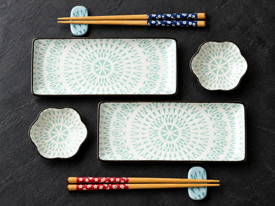 A set of Japanese-style ceramic dinnerware and chopsticks arranged on a dark slate background. The set includes two rectangular platters with a mint green radial pattern on a white glaze, two small flower-shaped bowls with matching patterns, and four pairs of wooden chopsticks with different decorative bands (blue with white flowers, red with white flowers, and light blue with white flowers).