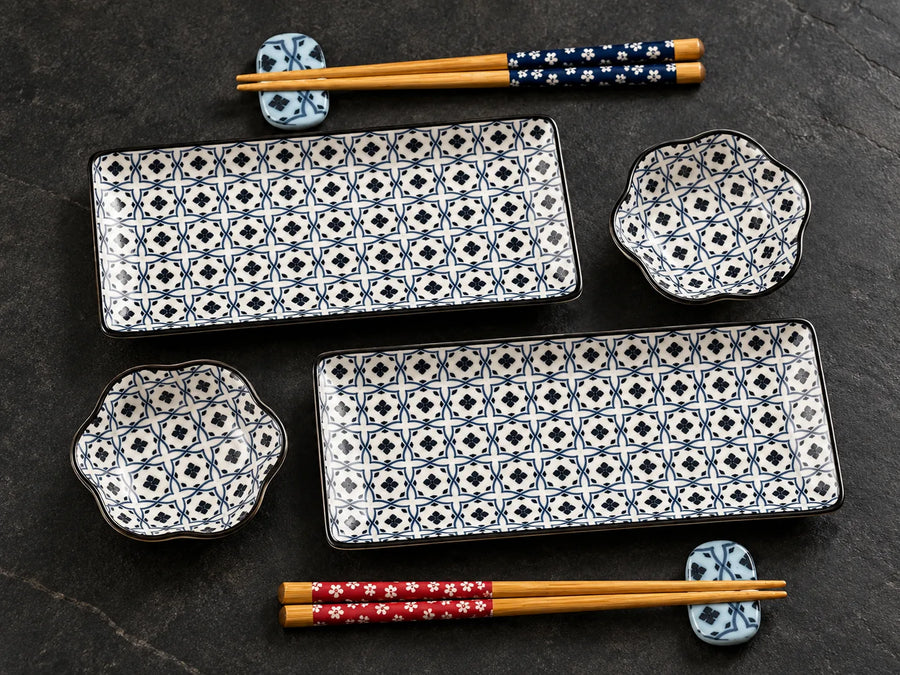 A set of ceramic tableware on a dark textured surface, including two rectangular plates with blue and white geometric patterns, two scalloped bowls with matching patterns, and a pair of wooden chopsticks with blue floral wrapping resting on a blue patterned chopstick holder.