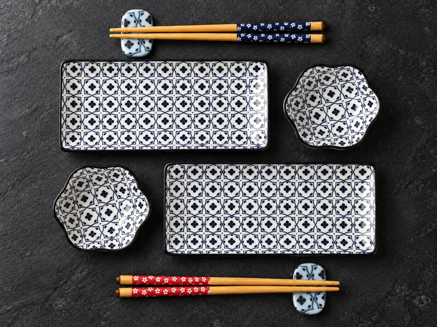 A set of ceramic tableware with a blue and white geometric pattern, including two rectangular platters, two scalloped bowls, and a pair of chopsticks with a blue floral wrap, arranged on a dark textured background.