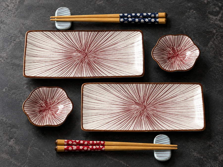 A set of Japanese-style ceramic tableware arranged on a dark textured surface, including two rectangular plates, two flower-shaped bowls, and two pairs of chopsticks with decorative fabric wraps.