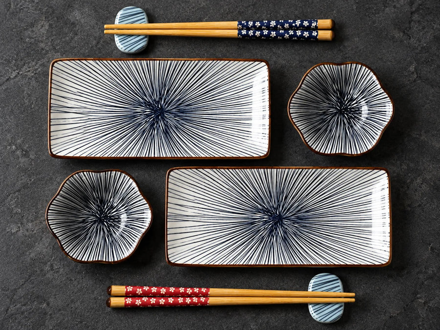 A set of ceramic tableware on a dark textured background, including two rectangular plates with blue radial line patterns, two flower-shaped bowls, and two pairs of wooden chopsticks with decorative fabric wraps.