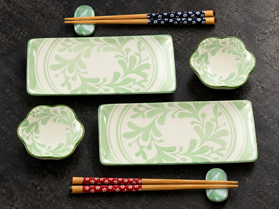 A set of green and white ceramic tableware with floral leaf patterns, including two rectangular serving platters, two small flower-shaped bowls, and four pairs of wooden chopsticks in decorative bands, arranged on a dark textured surface.