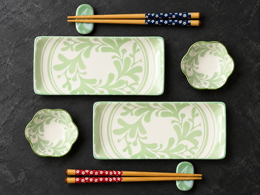 A set of green and white ceramic tableware with a leaf pattern, including two rectangular serving platters, two small scalloped bowls, and four pairs of chopsticks on matching green stands, arranged on a dark textured background.