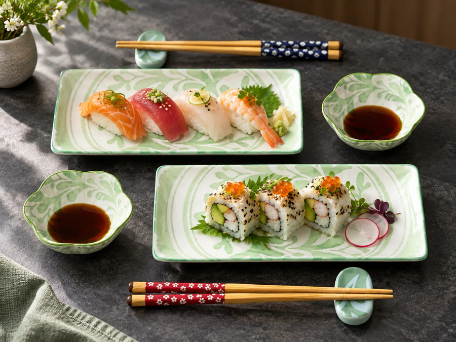 A styled tabletop arrangement of sushi dishes on green-and-white patterned rectangular plates, with small bowls of soy sauce, chopsticks, and a ceramic vase of flowers on a dark gray surface.