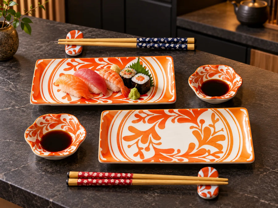 A set of orange and white ceramic sushi serving pieces on a dark stone countertop, including two rectangular plates with floral patterns, two small bowls of soy sauce, and two pairs of chopsticks with patterned fabric sleeves.