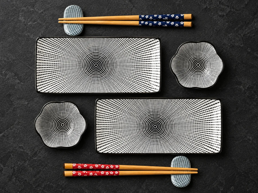 A flat-lay view of a ceramic dinnerware set on a dark textured background, featuring two rectangular plates with concentric circle patterns, two flower-shaped bowls, and four pairs of wooden chopsticks with decorative fabric bands.