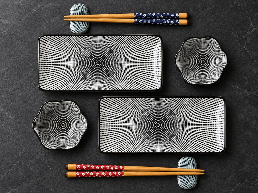 A set of black and white patterned ceramic tableware, including two rectangular plates, two flower-shaped bowls, and four pairs of chopsticks with decorative handles, arranged on a dark textured background.
