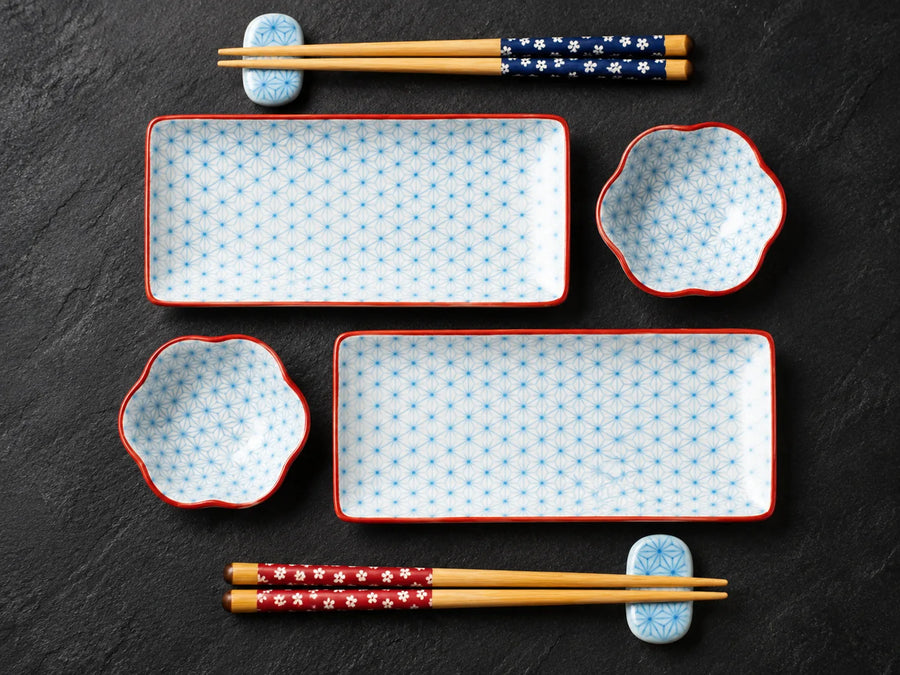 A set of white ceramic dinnerware with a light blue geometric pattern and red rims, including two rectangular platters, two flower-shaped bowls, and two pairs of chopsticks with blue and red floral wrapped handles, arranged on a dark slate background.