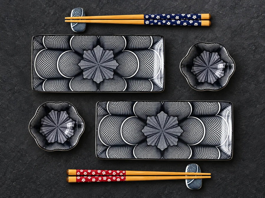 A set of black and gray ceramic Japanese-style dinnerware with floral patterns, including two rectangular plates, two small bowls, and four pairs of chopsticks with different patterned handles, arranged on a dark textured background.