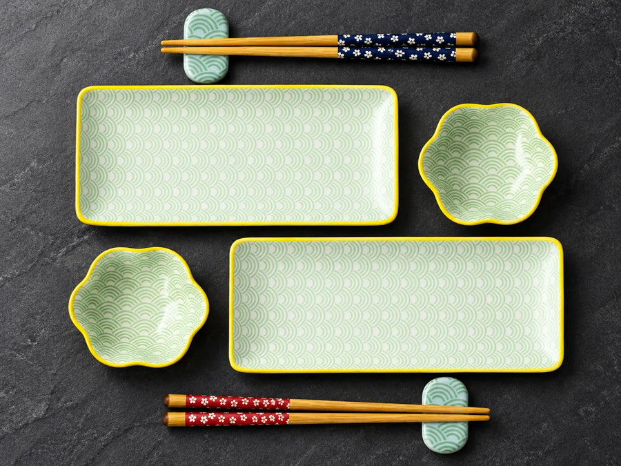 Japanese-style ceramic dinnerware set on a dark slate background, featuring two rectangular plates with yellow borders and scalloped wave-pattern interiors, two matching flower-shaped bowls, and four pairs of chopsticks with floral-patterned bands.