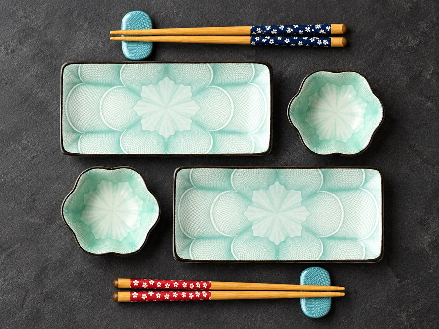 A set of Japanese-style ceramic tableware on a dark textured background, including two rectangular plates with embossed floral patterns, two small flower-shaped bowls, and four pairs of chopsticks in different color patterns.