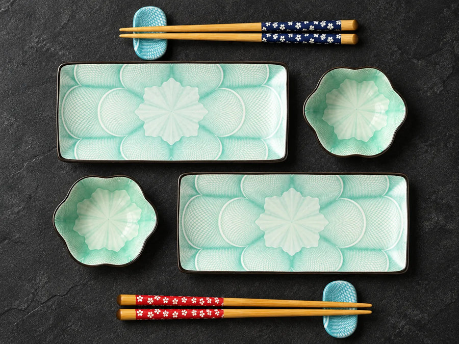 A Japanese-style ceramic dinnerware set arranged on a dark textured surface, featuring two rectangular plates with a white floral motif on a light aqua background, two matching flower-shaped bowls, and four pairs of wooden chopsticks with decorative floral bands in blue, red, and white.