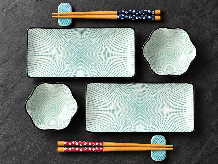 A set of ceramic dinnerware and chopsticks arranged on a dark slate background. The set includes two rectangular plates with radiating concentric dot patterns, two flower-shaped bowls with similar patterns, and a pair of wooden chopsticks with navy and red floral-patterned handles resting on matching light blue chopstick rests.