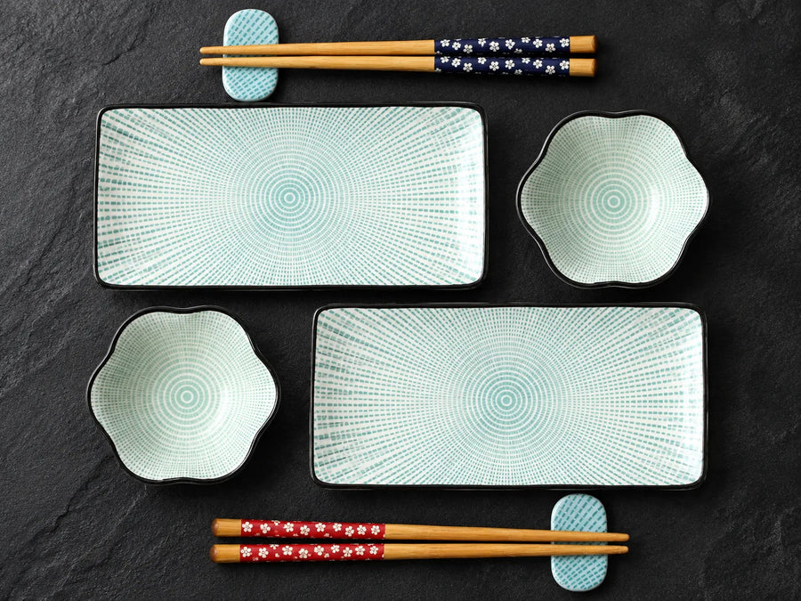 A flat-lay view of a Japanese-style dinnerware set on a dark slate background, including two rectangular plates with concentric circular patterns, two flower-shaped bowls, and four pairs of chopsticks with decorative bands.