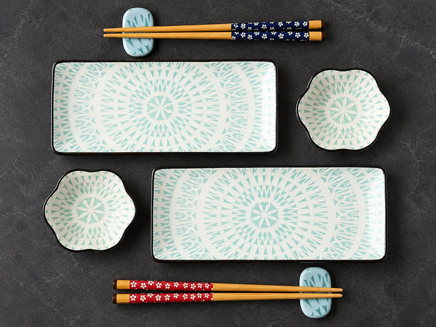 A Japanese-style dinnerware set arranged on a dark textured surface, including two rectangular platters with scalloped edges, two small flower-shaped bowls, and two pairs of chopsticks on decorative holders. The dinnerware has a white base with a light aqua circular sunburst pattern, while the chopsticks feature floral patterns in navy, red, and white.